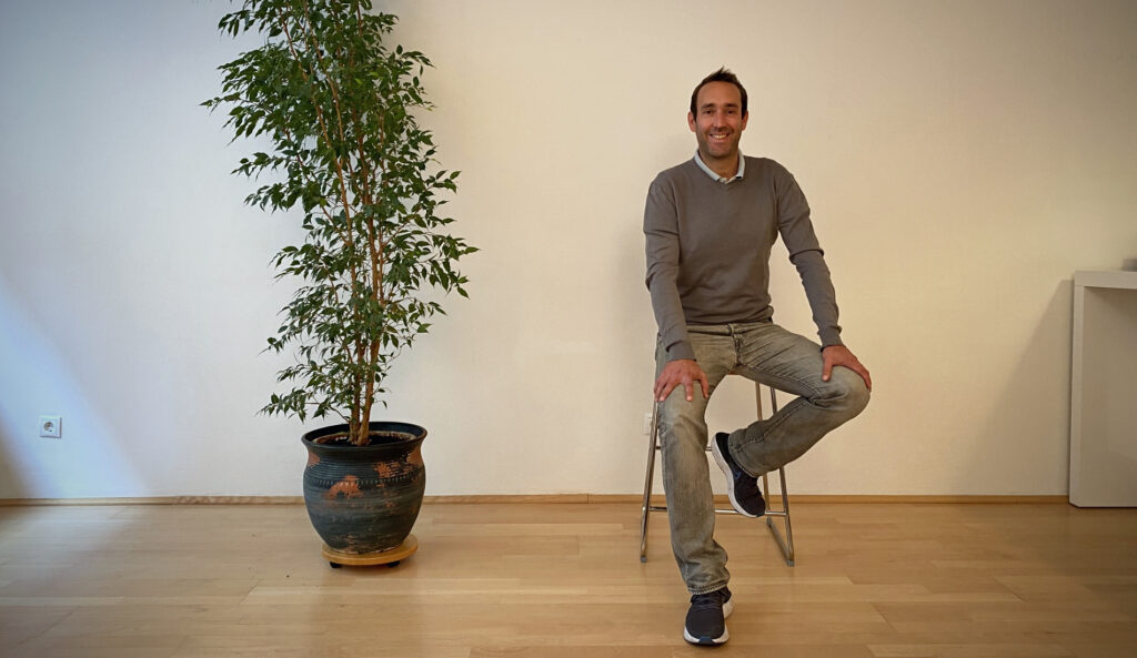 Matthias Tschannett, a psychotherapist in 1080 Vienna, sits smiling on a high chair next to a tall houseplant in his practice.